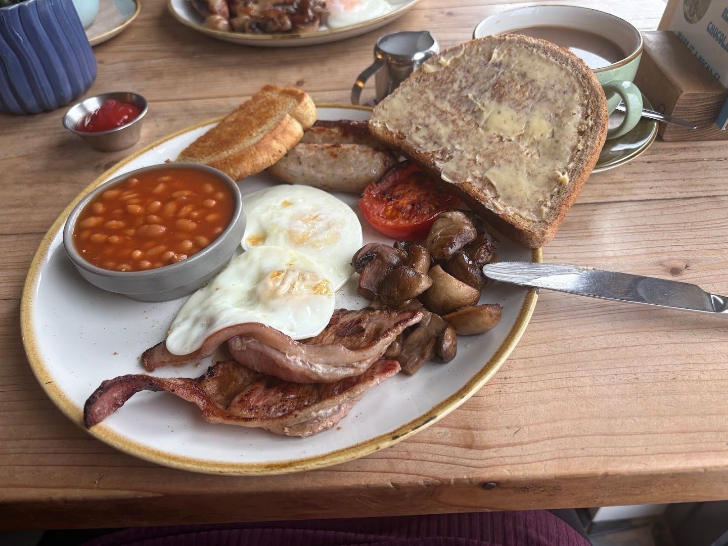 A cooked breakfast on a wooden table: bacon, fried eggs, mushrooms, tomato, sausage, beans and buttered toast