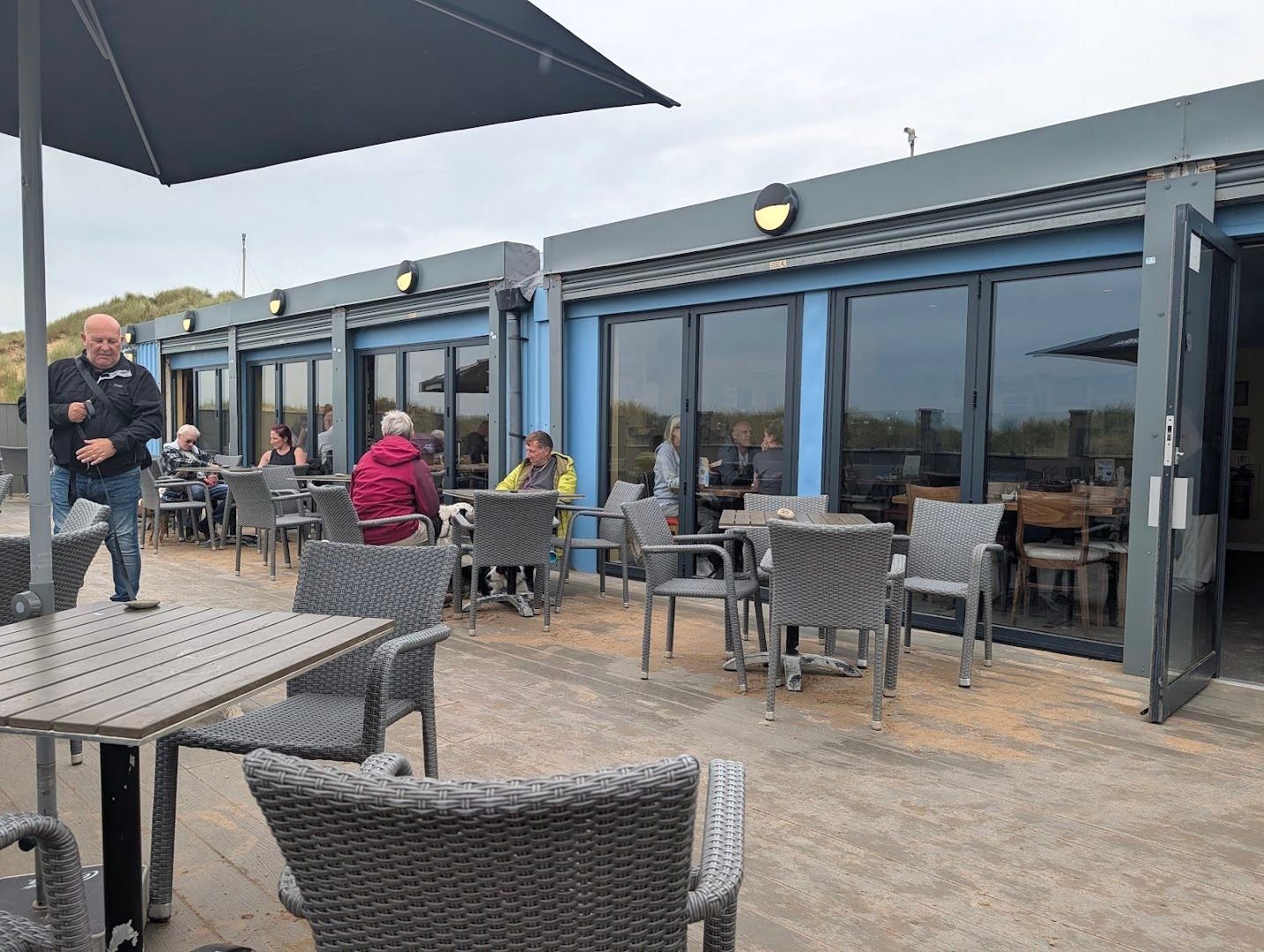 The café's decking under a grey sky: grey wicker chairs and tables, a parasol, and the long glass front of the building with blue frames