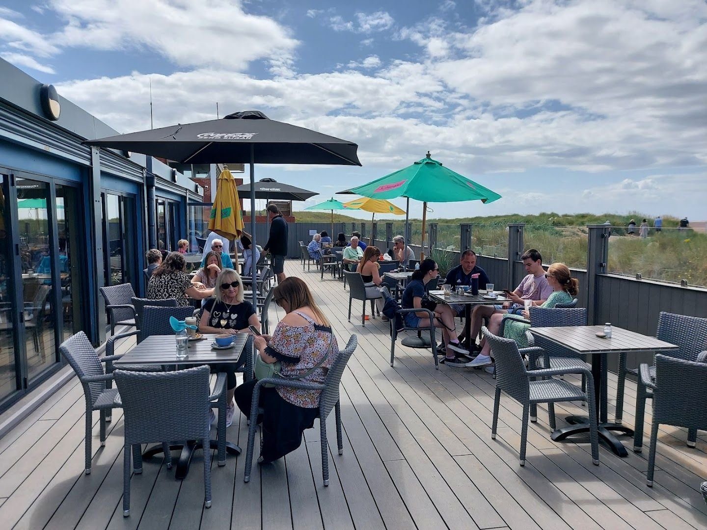 The deck on a sunny day, busy with people at grey tables under black and green parasols, with the dunes behind
