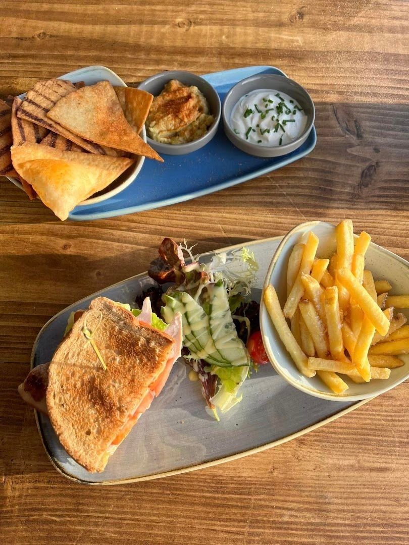 Lunch on a wooden table: a toasted sandwich with salad and chips, and a plate of tortilla chips with dips